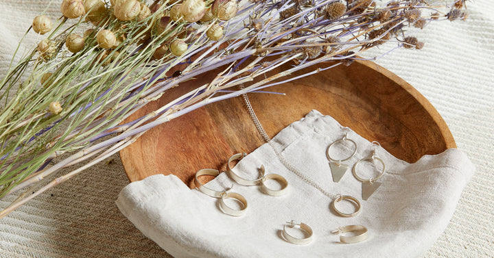 A selection of silver and brass jewellery laid on a linen napkin in a wooden bowl, with a sheaf of dried cotton stems 